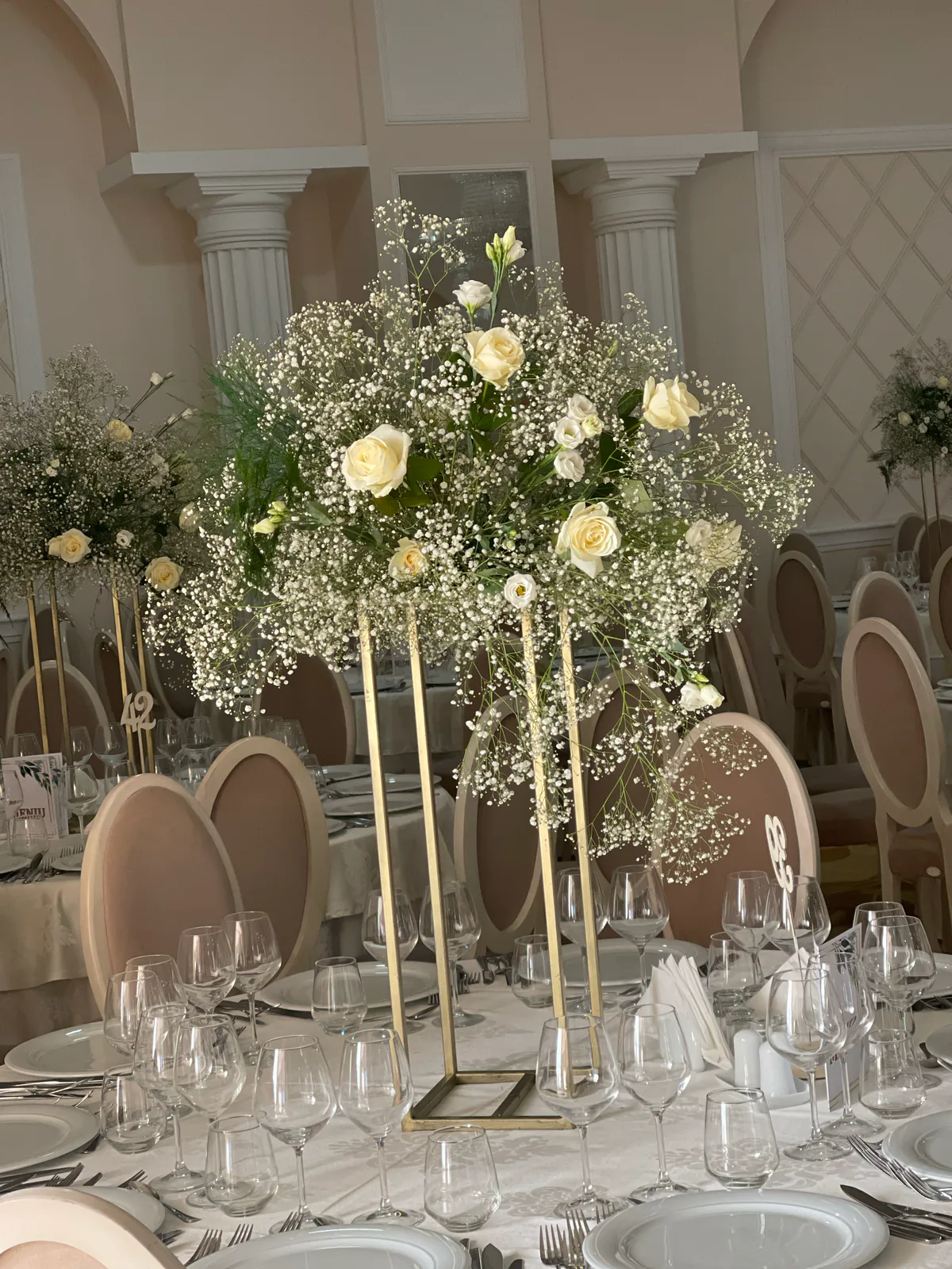Centru de masă înalt din trandafiri albi și gypsophila, pe suport auriu, într-o sală de evenimente