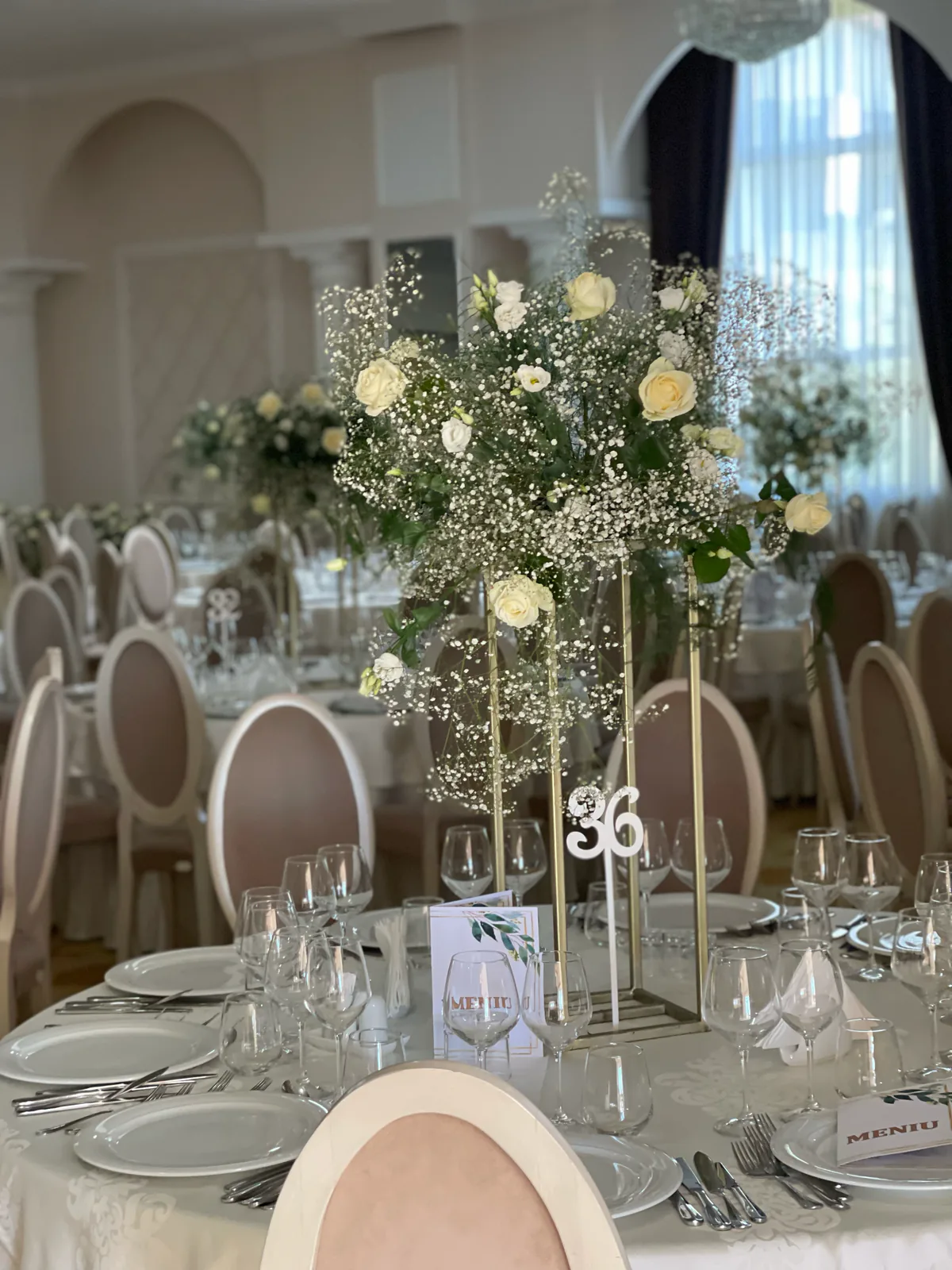 Sală de nuntă decorată cu aranjamente înalte din trandafiri albi și gypsophila pe mesele invitaților