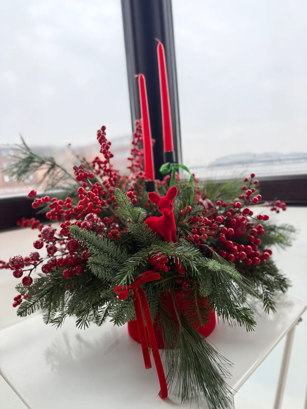 Aranjament de Crăciun cu ilex roșu, cetină și lumânări roșii, așezat la fereastră