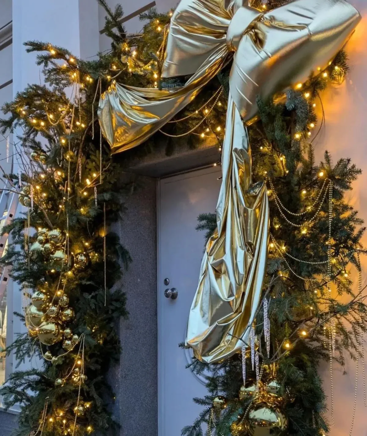 Intrare decorată de Crăciun cu ghirlandă de brad, lumini calde și fundă aurie mare